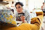 Young woman with headphones and credit card online shopping at laptop on living room sofa