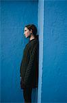 Portrait of teenage girl with brown hair in ponytail wearing black knitted jumper, leaning against blue wall.