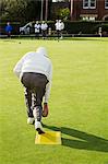 A lawn bowls player standing on a small yellow mat preparing to deliver a bowl down the green, the smooth grass playing surface.