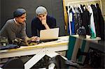 Two men working at a laptop, sharing the screen. A tailoring workshop