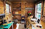 A designer seated in his leatherwork workshop, at a desk using a laptop. Woodburning stove with a glowing fire lit.