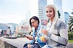 Young couple with takeaway juice and smartphone sitting on wall in city