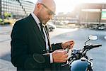 Mature businessman outdoors, standing beside motorcycle, holding eyeglasses