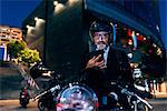 Mature businessman outdoors at night, sitting on motorcycle, using smartphone