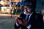 Mature businessman outdoors at night, wearing motorcycle helmet, using smartphone