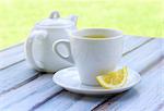 Green tea in a white cup, a slice of lemon on a saucer and a teapot on a wooden table.