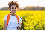 Outdoor portrait in golden evening sunshine of beautiful happy mixed race African American girl teenager female young woman smiling laughing with perfect teeth hiking with orange rucksack and water bottle in field of yellow flowers