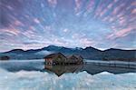 Pink clouds at sunset and wooden huts are reflected in the clear water of Kochelsee, Schlehdorf, Bavaria, Germany, Europe