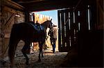 Rear view of cowboy leading saddled horse out of stable.