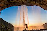 Rear view of man standing underneath natural arch behind waterfall at sunset.