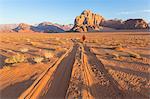 Rear view of man walking in desert landscape with tire tracks leading to distant mountain.