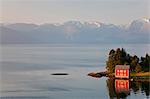 Traditional red wooden house standing on sea shore, snow capped mountains in the distance.