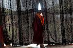 Rear view of young monk wearing red robe looking through black curtains at a monastery.