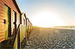 View along a row of colourful wooden beach huts on a long sandy beach at sunrise.