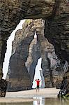 Rear view of man wearing red top standing underneath natural rock arch on sandy beach.