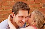 Girl whispering in father's ear by brick wall, head and shoulder