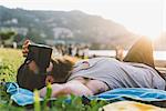 Young male hipster lying on grass looking at virtual reality headset, Lake Como, Lombardy, Italy