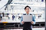 Businessman wearing blue shirt and vest standing on train station platform, holding laptop, looking at camera.