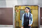 Businessman wearing blue shirt and vest standing on train station platform, holding laptop.