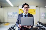 Businessman wearing suit and glasses standing at train station, holding laptop.