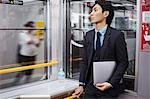 Businessman wearing suit standing on a commuter train, holding laptop.