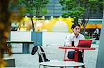 Businessman wearing white shirt sitting outdoors at red table, working on laptop.