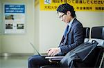 Businessman wearing suit and glasses sitting at train station, working on laptop.