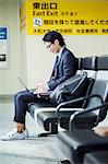 Businessman wearing suit and glasses sitting at train station, working on laptop.