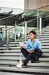 Businessman wearing blue shirt sitting outdoors on steps, holding digital tablet and papers.