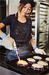 Woman working in a bakery, wearing oven gloves, using blowtorch, melting cheese on sandwiches.