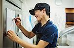 Man working in a bakery, wearing baseball cap and apron, writing note on small whiteboard, using phone and smiling.