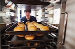 Man working in a bakery, placing large trays with freshly baked rolls on a trolley.