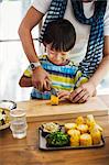 Man and boy standing at a table, preparing corn on the cob, smiling.