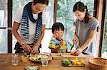 Man, woman and boy standing at a table, preparing corn on the cob, smiling.