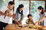 Two men, woman and boy gathered around a table, preparing corn on the cob, one man holding cat, smiling.