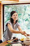 Smiling woman with black hair standing at a table with bowls of food, holding wooden ladle.