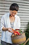 Smiling woman standing outdoors, holding basket and freshly picked tomatoes.