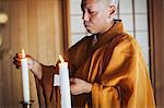 Side view of Buddhist monk with shaved head wearing golden robe kneeling indoors in a temple, lighting white candle.