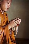 Side view of Buddhist monk with shaved head wearing golden robe kneeling indoors in a temple, holding mala, praying.