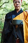 Buddhist monk with shaved head wearing black and yellow robe, standing outdoors, holding singing bowl.