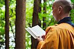 Side view of Buddhist monk with shaved head wearing black and yellow robe, standing outdoors, holding prayer text.