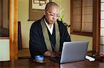 Buddhist monk with shaved head wearing black robe sitting indoors at a table, using laptop computer.