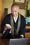 Buddhist monk with shaved head wearing black robe sitting indoors at a table in front of laptop computer, using mobile phone.