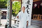 Buddhist monk wearing white robe and cap standing outside a temple, smiling at camera.