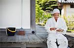 Buddhist monk wearing white robe and cap sitting on wooden floor outside a temple, using mobile phone.