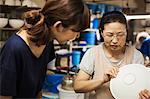 Two women standing in a Japanese porcelain workshop, inspecting a white bowl.