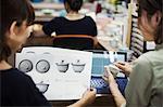 Two women sitting in a workshop at laptop, holding drawing of  Japanese porcelain cups and lids.