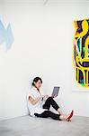 Woman with short black hair wearing white shirt, black trousers and red high heel shoes sitting on floor i art gallery, holding laptop.