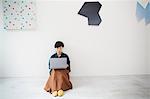 Woman with short black hair wearing green shirt sitting on floor in art gallery, balancing laptop on her knees.