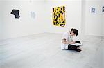 Woman with black hair wearing white shirt sitting on floor in art gallery with pen and paper.